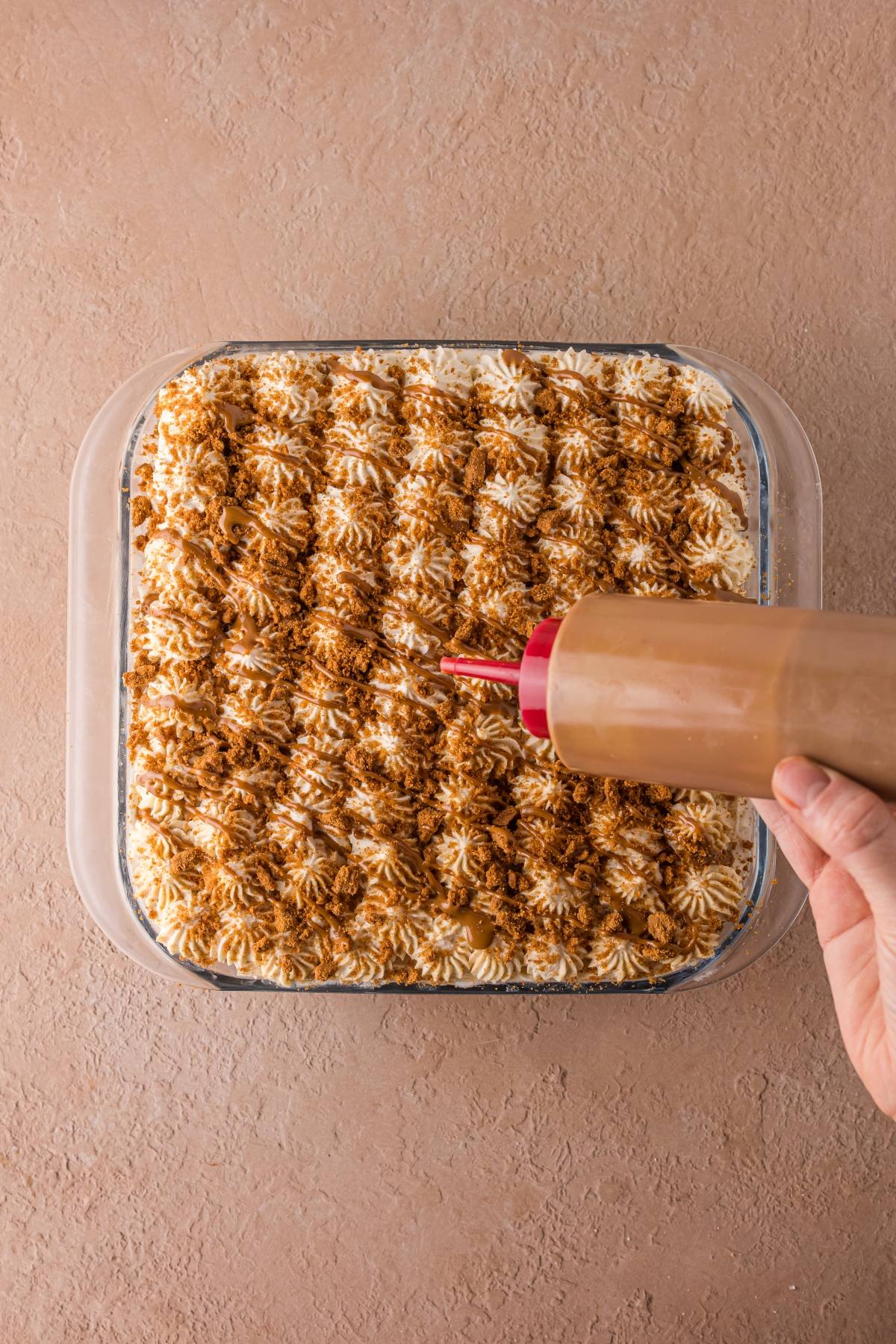 A hand drizzles sauce over a square dessert topped with piped cream and brown crumbs.