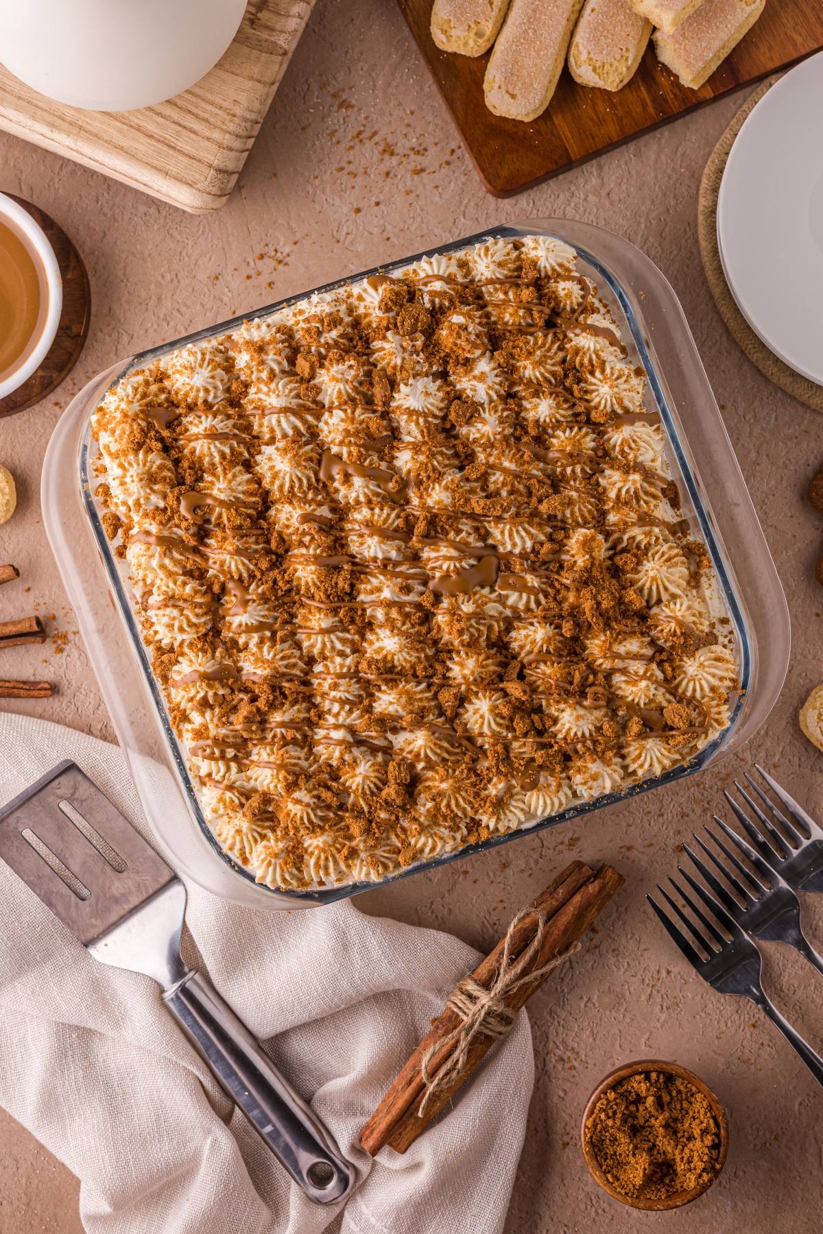 A square Biscoff tiramisu topped with piped cream and crumbled cookies, surrounded by utensils and cinnamon sticks.