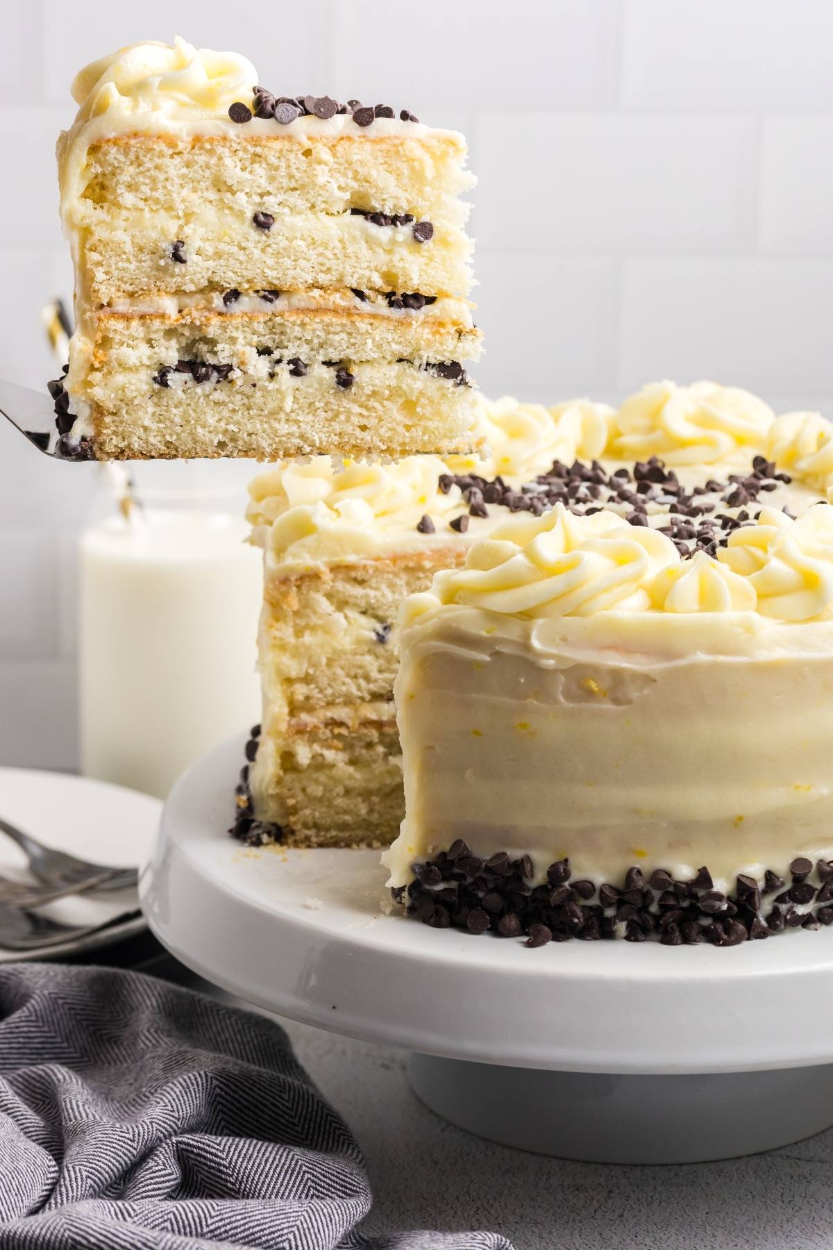 A slice of layered vanilla Cannoli cake  with chocolate chips is lifted from a frosted cake on a white stand.