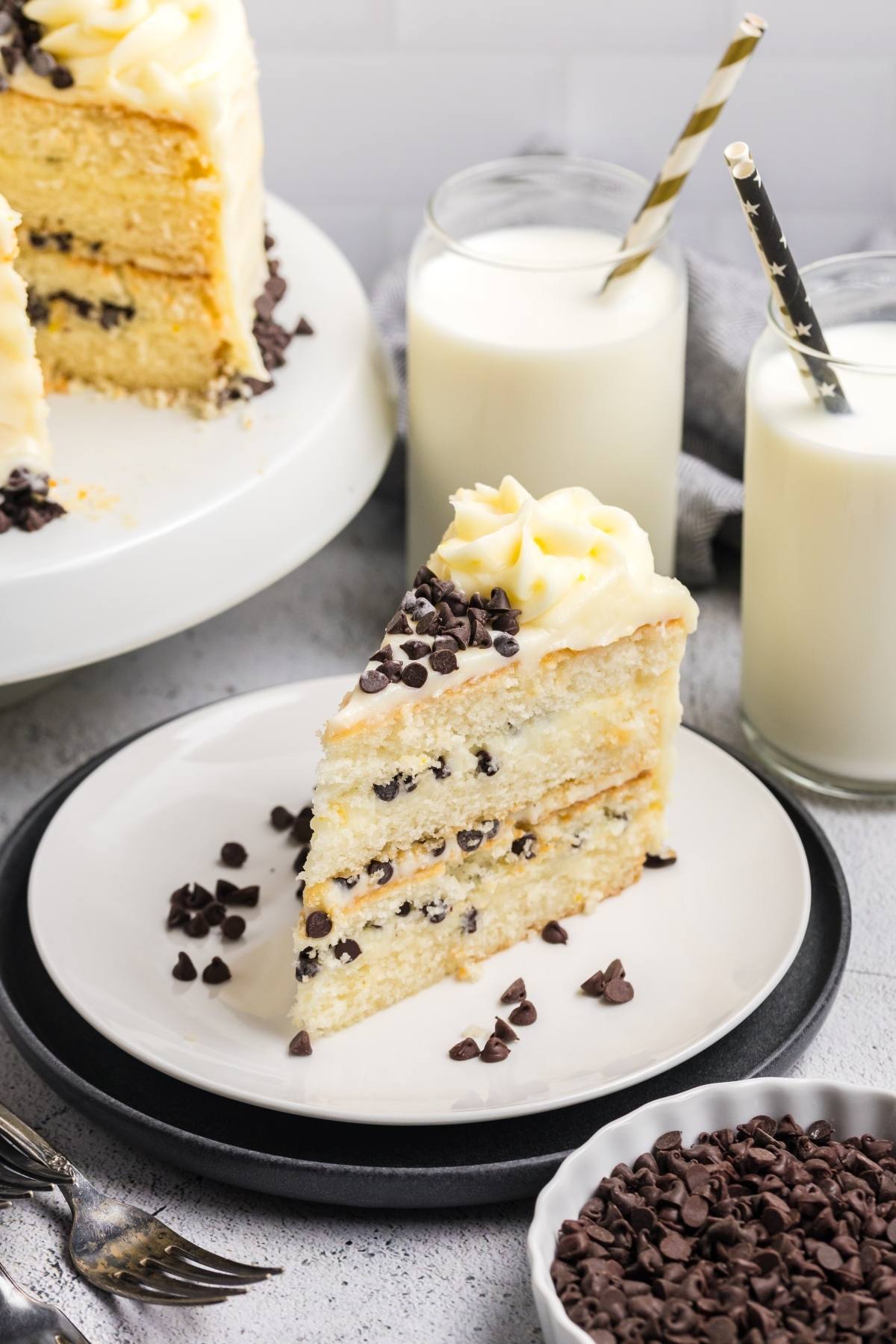 A slice of Cannoli cake with chocolate chips on a plate, two glasses of milk, and extra chocolate chips nearby.