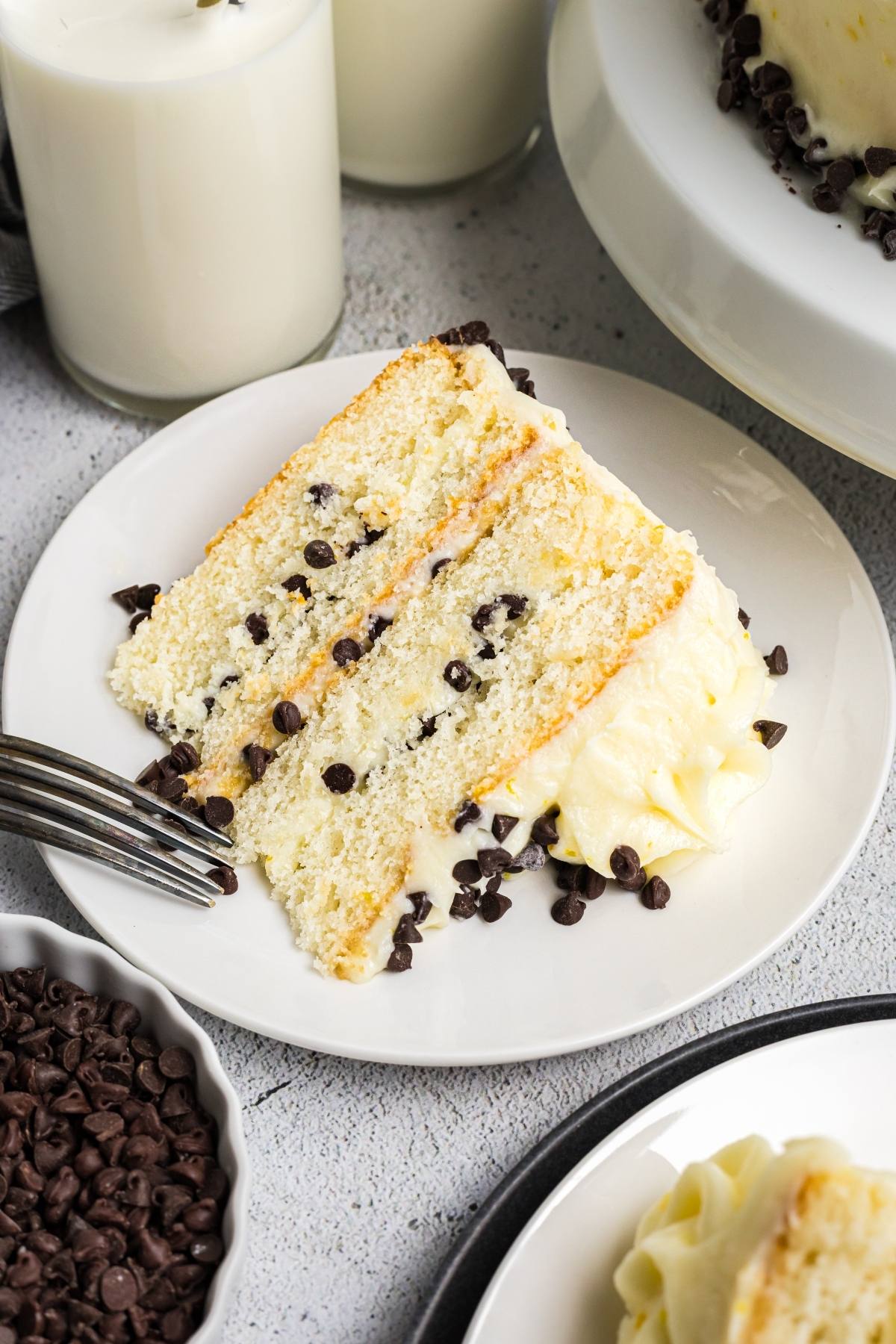 A slice of vanilla layer  Cannoli cake with chocolate chips and frosting on a white plate, next to a fork and milk.