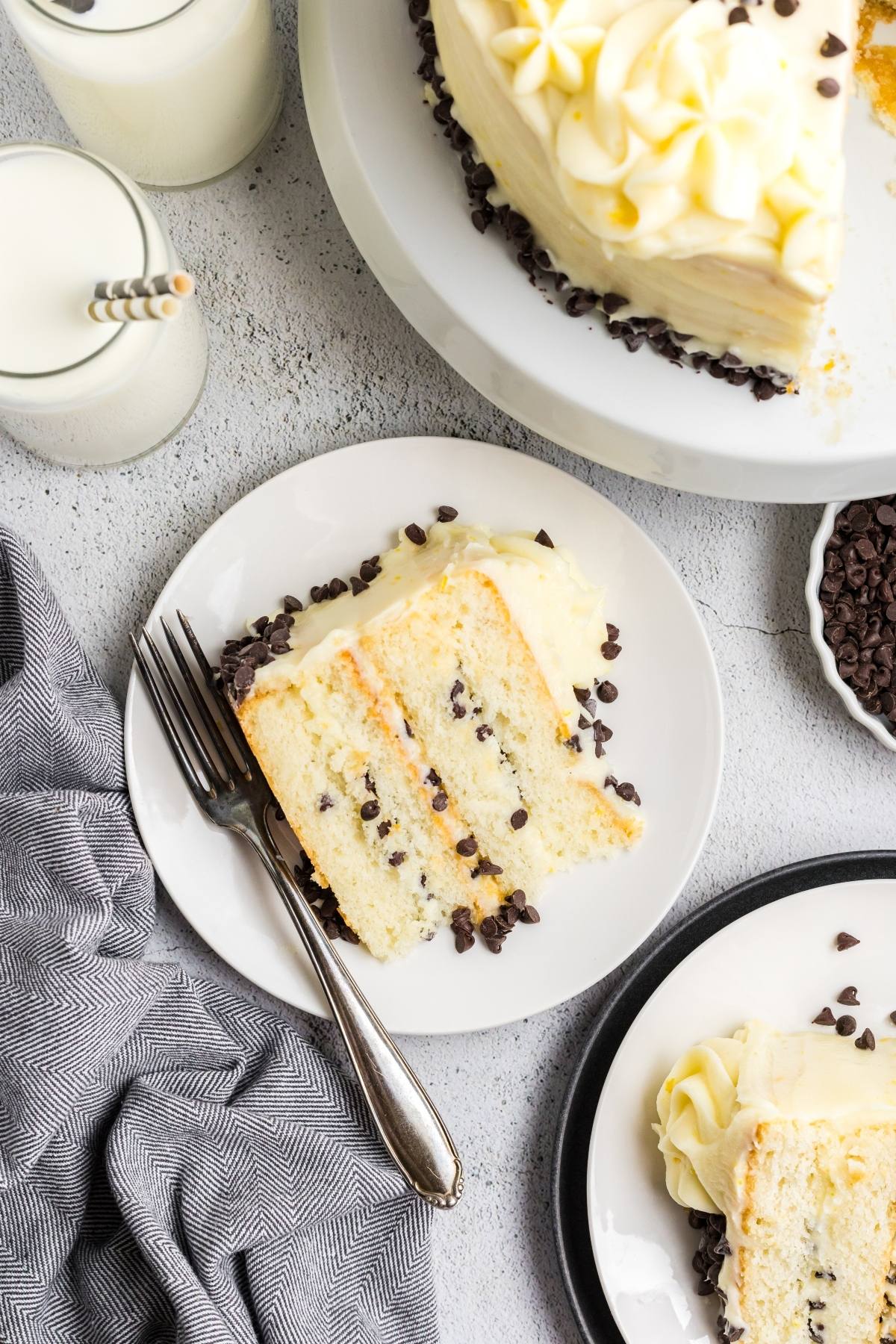 A slice of vanilla  Cannoli cake with chocolate chips on a plate, next to a fork, milk, and the rest of the cake.