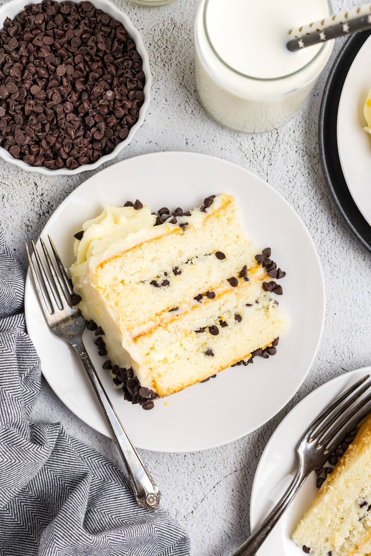 A slice of yellow  Cannoli cake with chocolate chips and white frosting on a plate, next to a fork and a bowl of chocolate chips.