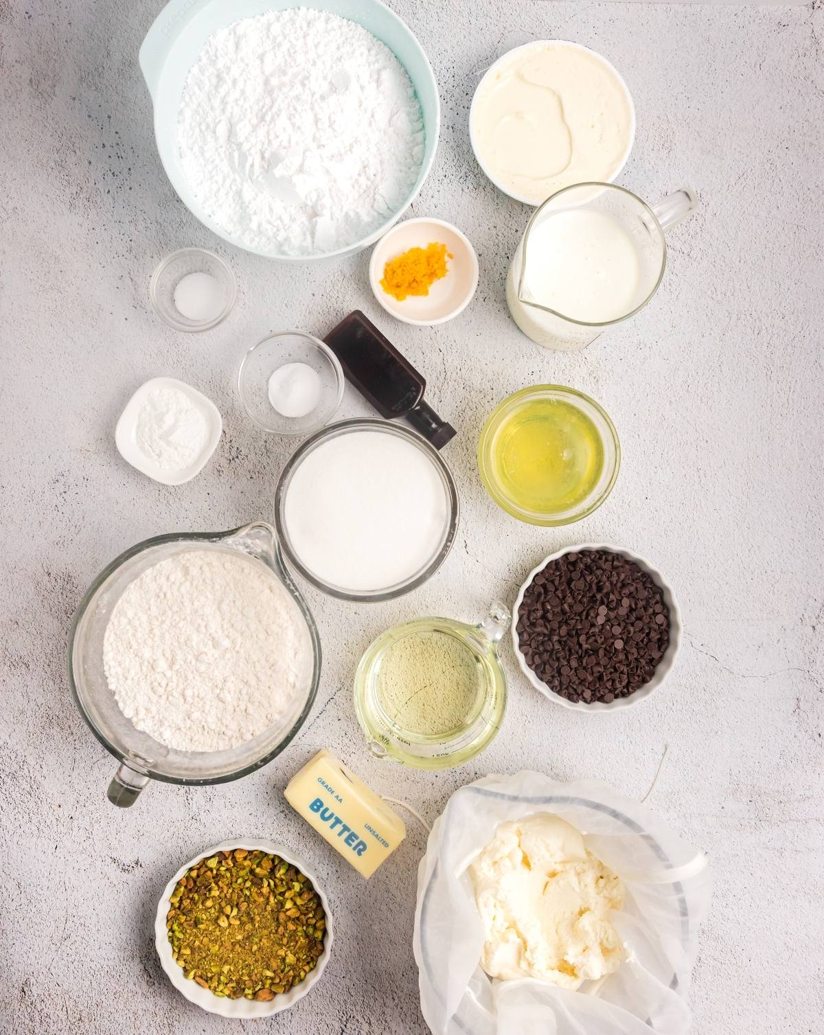 Baking ingredients including flour, sugar, chocolate chips, pistachios, butter, and bowls of liquids on a gray surface.