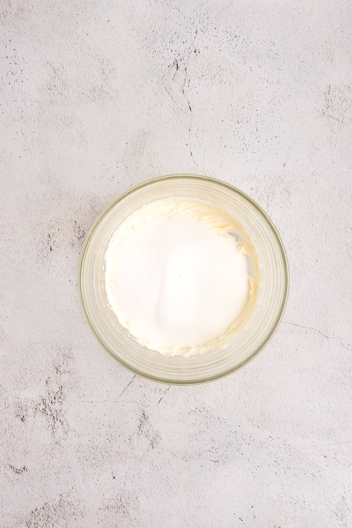 A glass bowl with white cream and sugar on a light textured surface.