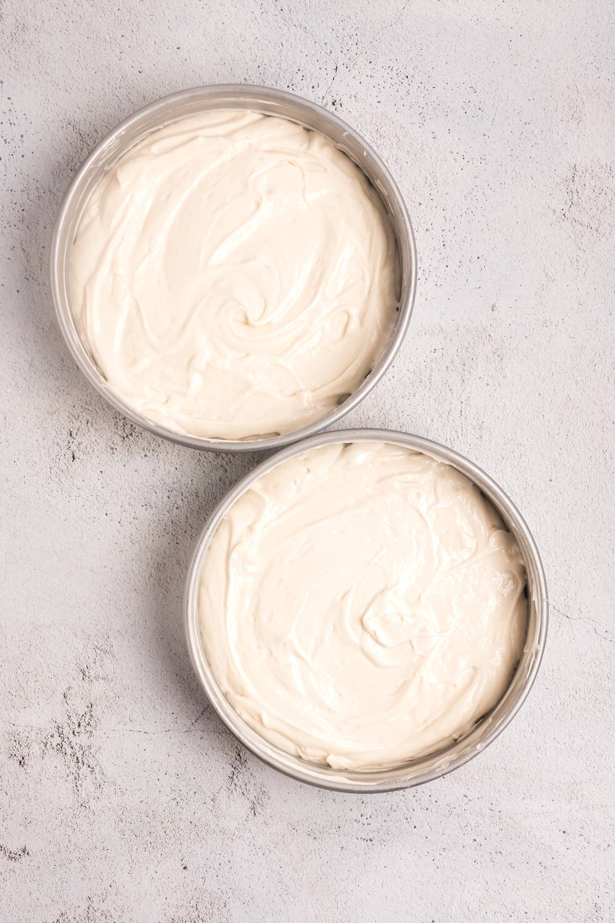 Two round cake pans filled with swirled, unbaked cake batter on a light textured surface.