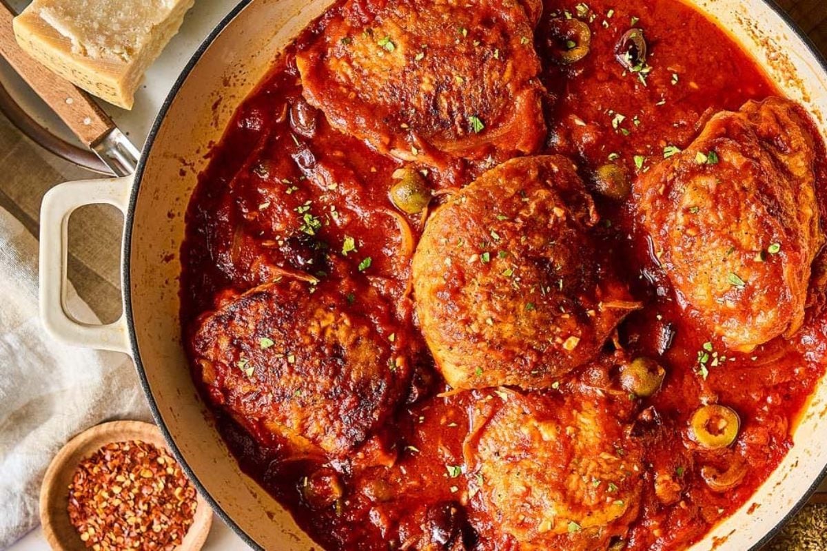 Chicken thighs in a rich tomato sauce with olives, served in a Dutch oven, garnished with herbs.