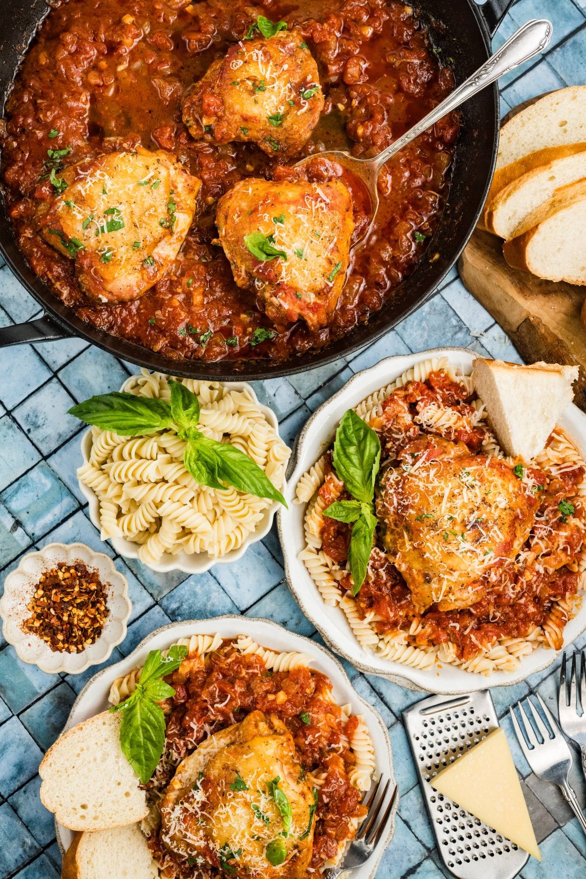 Two plates of pasta topped with Chicken fra diavolo in tomato sauce, bread slices, grated cheese, and a skillet of chicken.