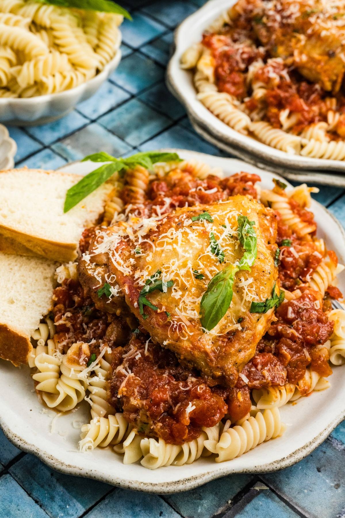 A plate of rotini pasta topped with tomato sauce, Chicken fra diavolo   grated cheese, basil, and two slices of bread.