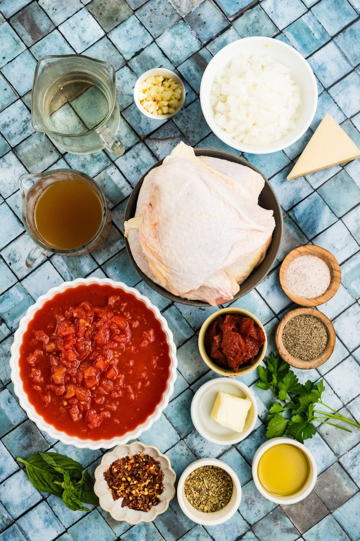 Top-down view of chicken, chopped veggies, herbs, spices, tomatoes, broth, oil, butter, and cheese on a blue tile surface.