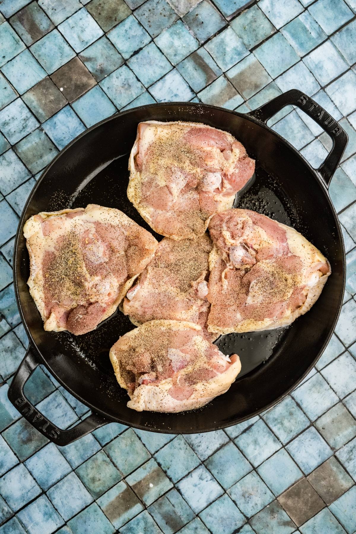 Raw, seasoned chicken thighs arranged in a round black pan on a blue tiled surface.