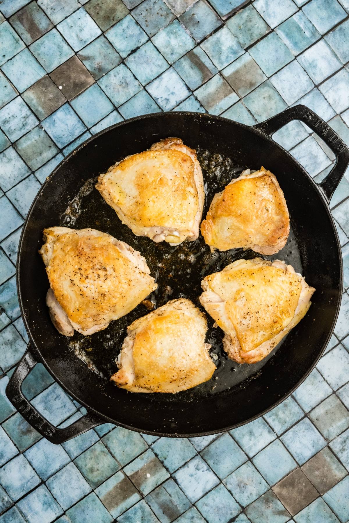 Five seasoned chicken thighs cooking in a black skillet on a blue tiled surface.