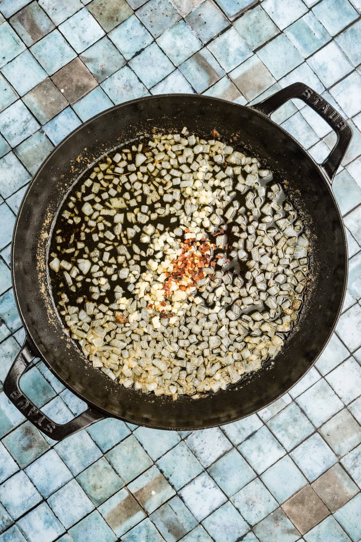 Chopped onions, garlic, and red pepper flakes cooking in oil in a large black pan on a tiled surface.