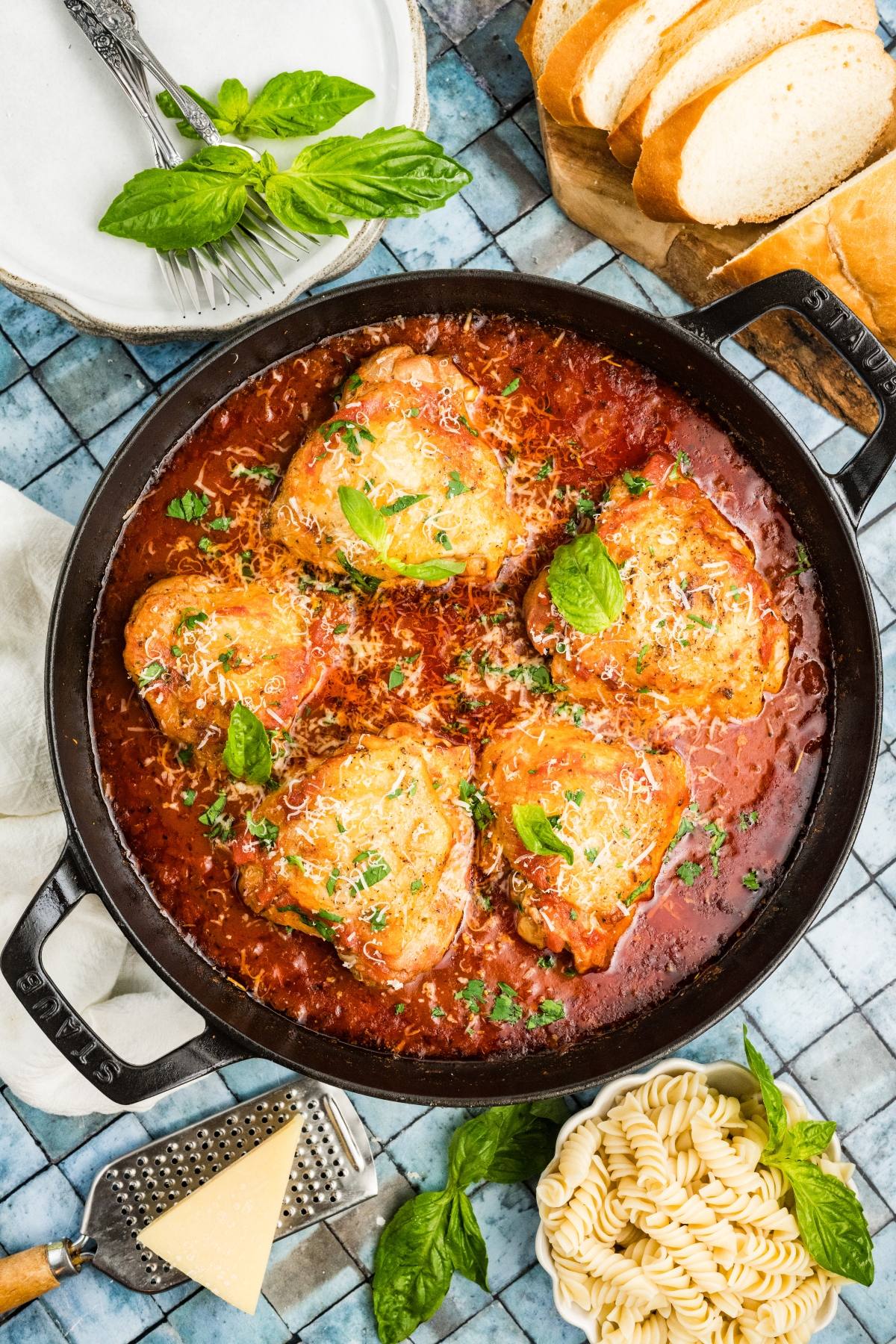 A skillet of Chicken fra diavolo parmesan in tomato sauce, garnished with basil, beside pasta, bread, and parmesan cheese.