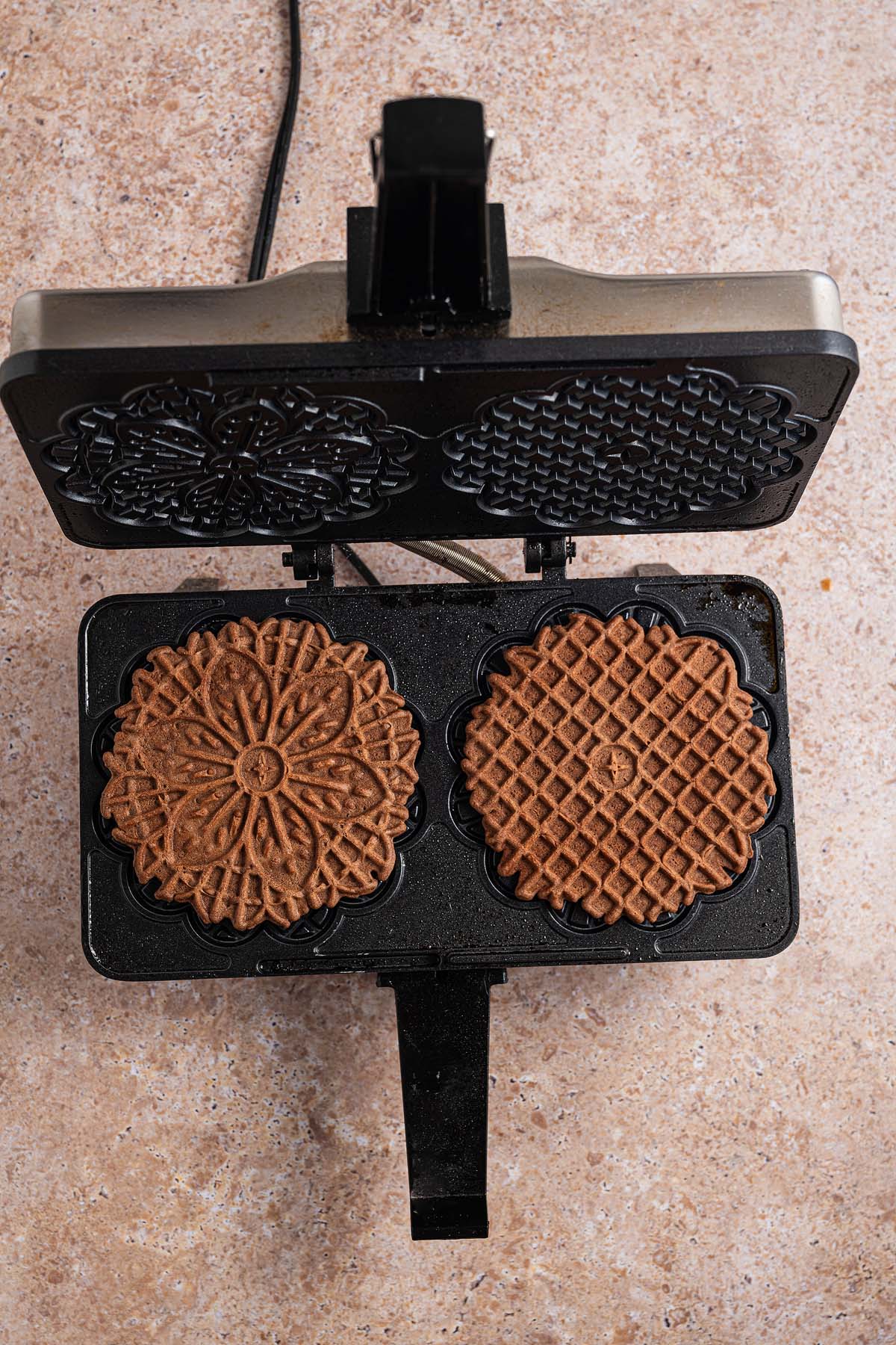 Two chocolate pizzelle cookies cook in a black pizzelle maker on a beige countertop, filling the air with the rich aroma of chocolate pizzelle.