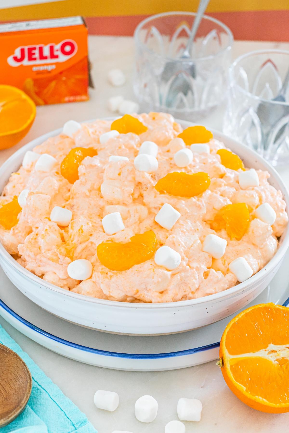 A bowl of orange Cottage cheese jello salad topped with mandarin slices and mini marshmallows, next to oranges and Jello box.