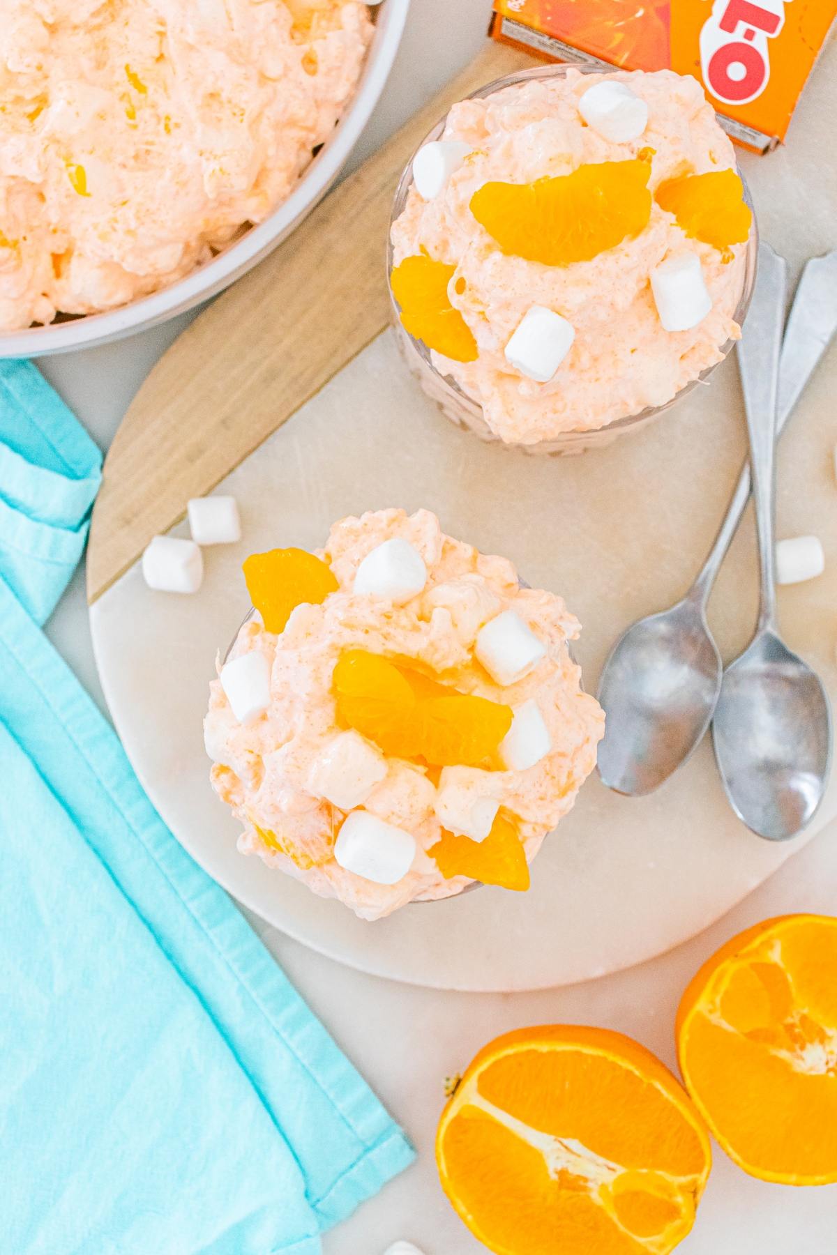 Two bowls of Cottage cheese jello salad with mini marshmallows and orange slices, next to spoons and fresh oranges.