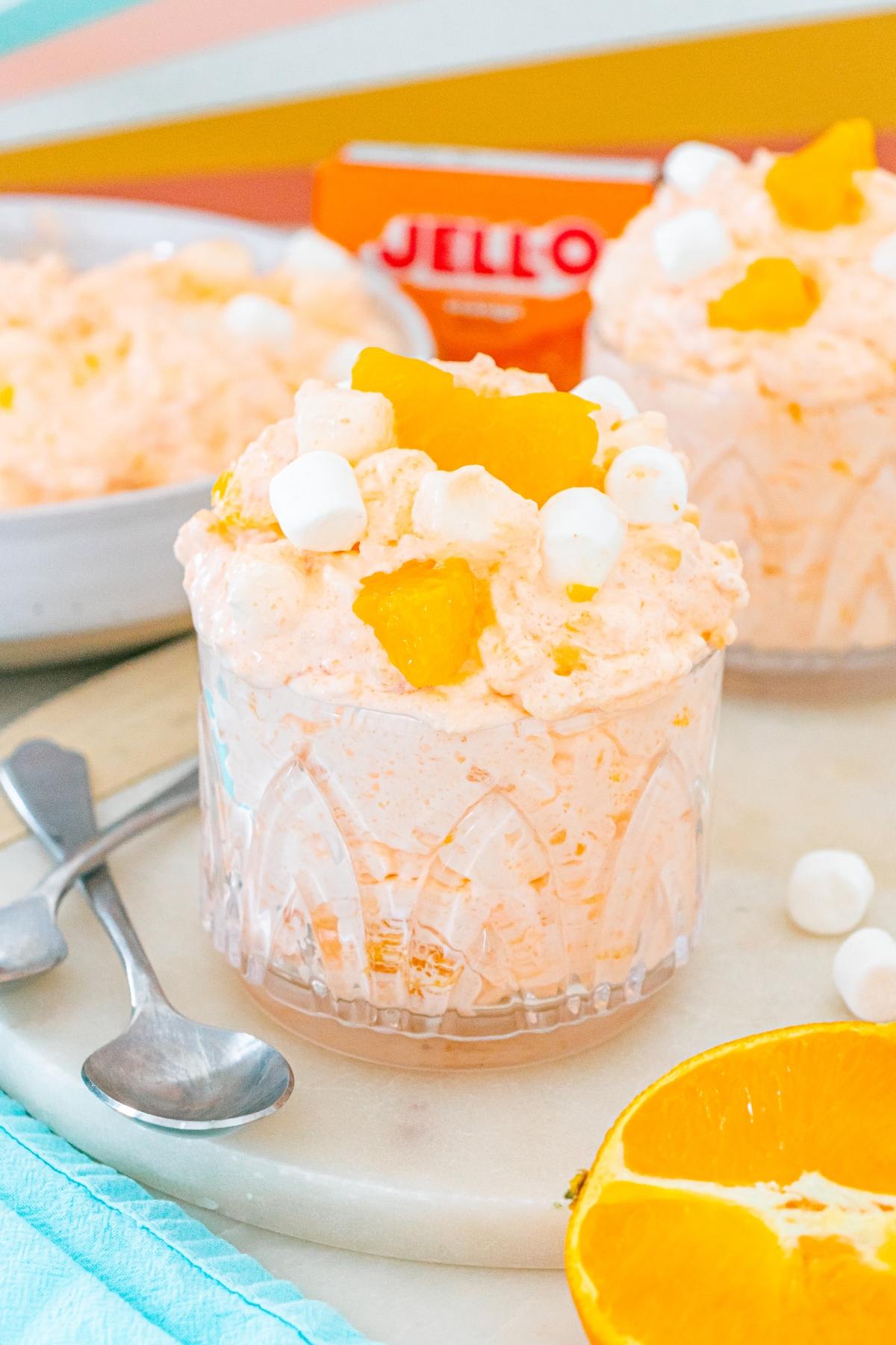 Glass of Cottage cheese jello salad topped with orange pieces and mini marshmallows, with spoons nearby.