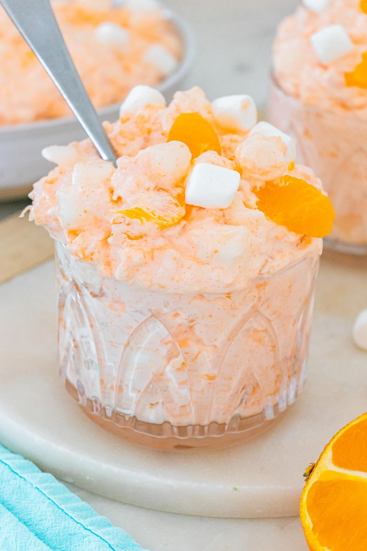 A glass of Cottage cheese jello salad creamy orange fluff salad topped with mandarin slices and mini marshmallows, with a spoon inside.