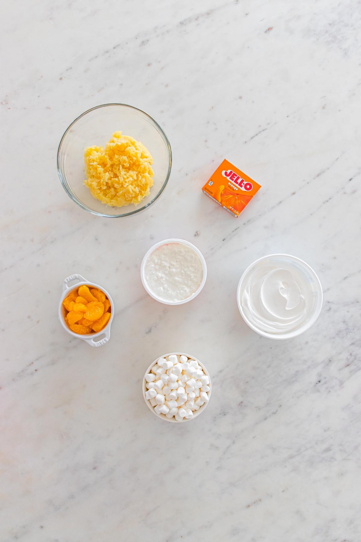 Five bowls with pineapple, whipped topping, marshmallows, oranges, cottage cheese, and a box of orange Jell-O.