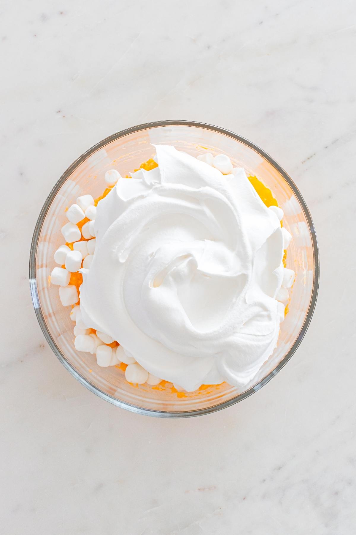 Glass bowl with mini marshmallows, orange mixture, and whipped topping on a light marble surface.