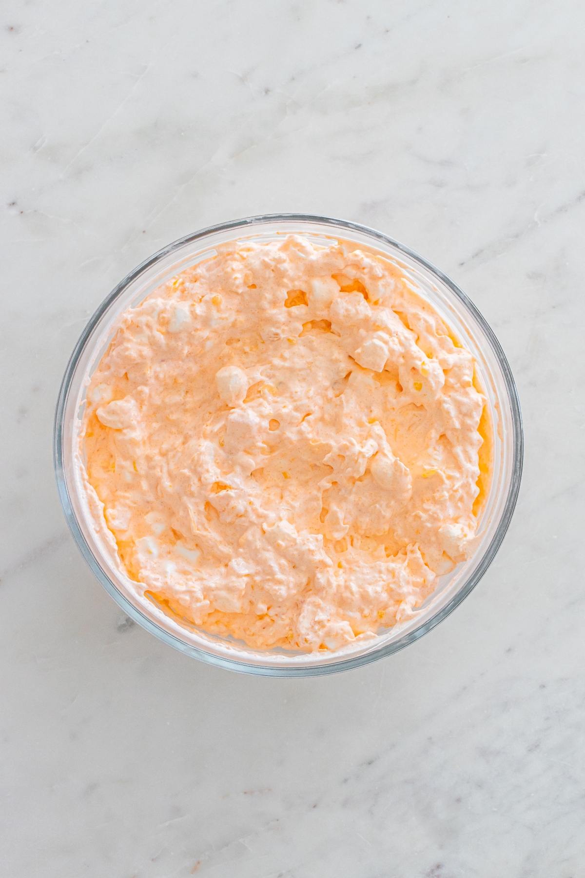 A glass bowl filled with light orange, fluffy whipped dessert on a white marble surface.