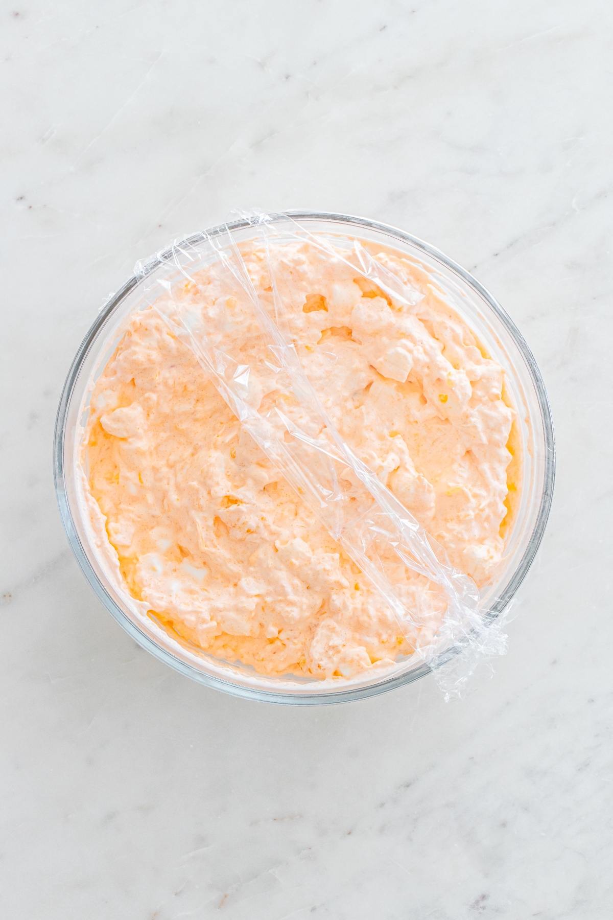 A glass bowl of orange-colored creamy dip covered with plastic wrap on a marble surface.