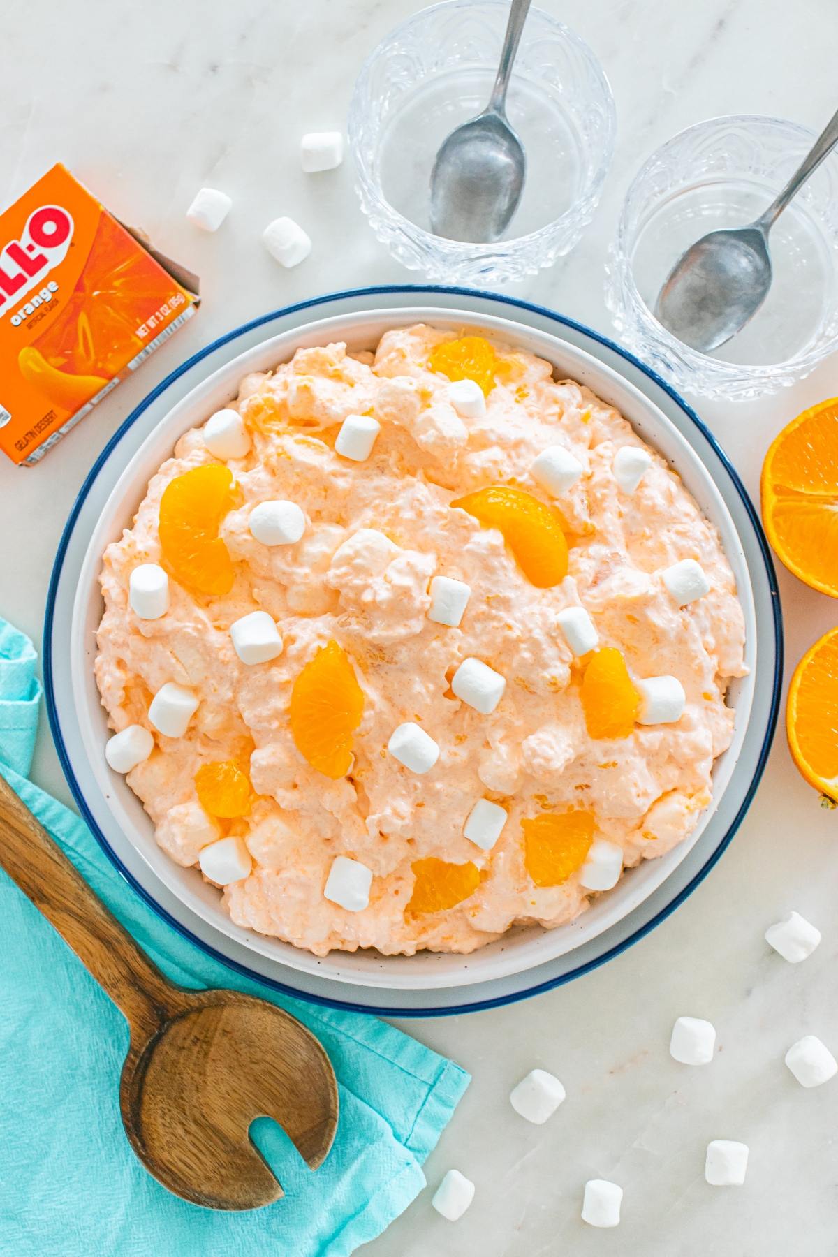 A bowl of Cottage cheese jello salad with marshmallows and orange slices, next to utensils, orange halves, and Jell-O box.