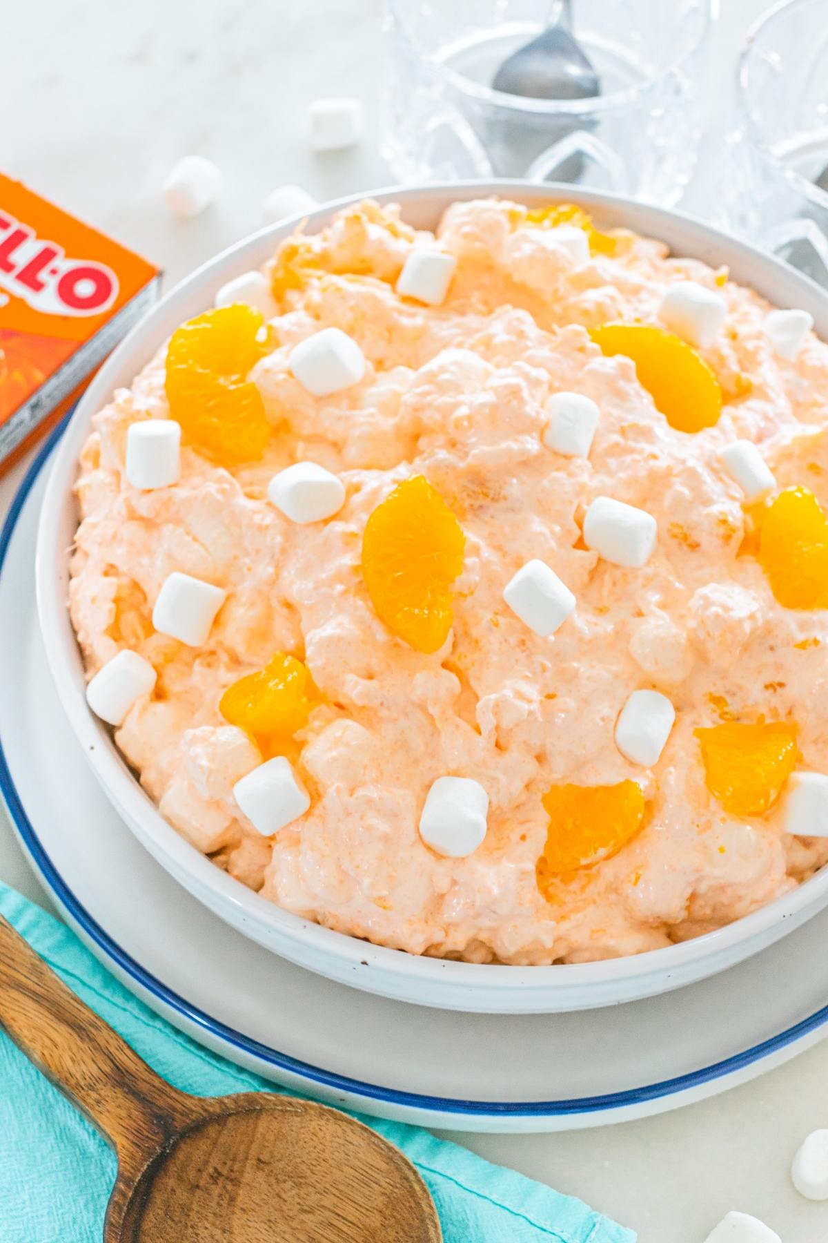 A bowl of Cottage cheese jello salad with mandarin slices and mini marshmallows on top, next to a wooden spoon.