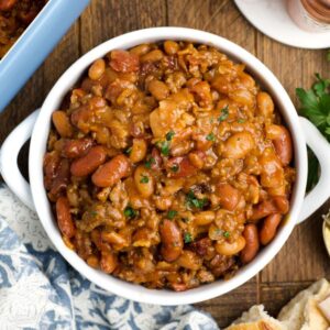 A bowl of cowboy baked beans with ground meat, garnished with herbs, sits on a wooden table alongside bread and utensils.