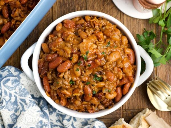 A bowl of cowboy baked beans with ground meat, garnished with herbs, sits on a wooden table alongside bread and utensils.