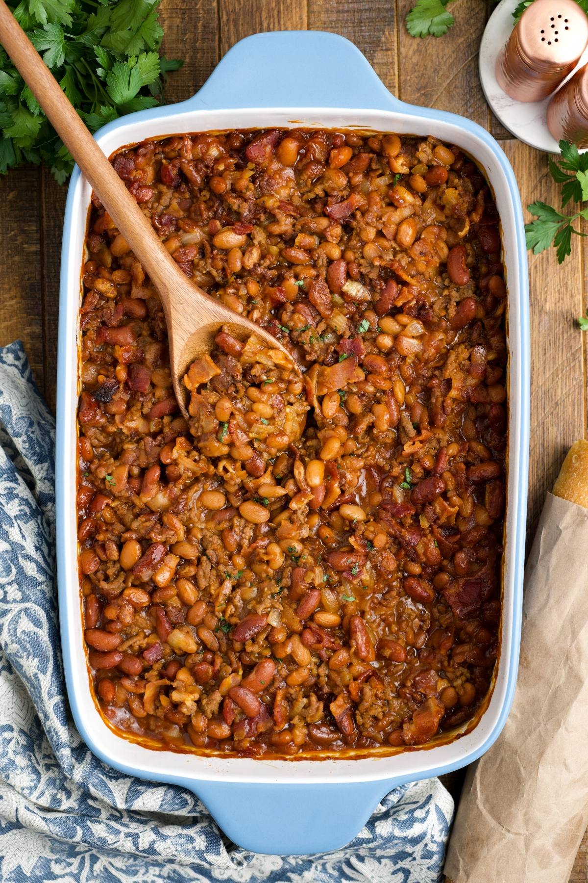 cowboy Baked beans in a blue casserole dish with a wooden spoon, on a wooden table with bread and herbs nearby.