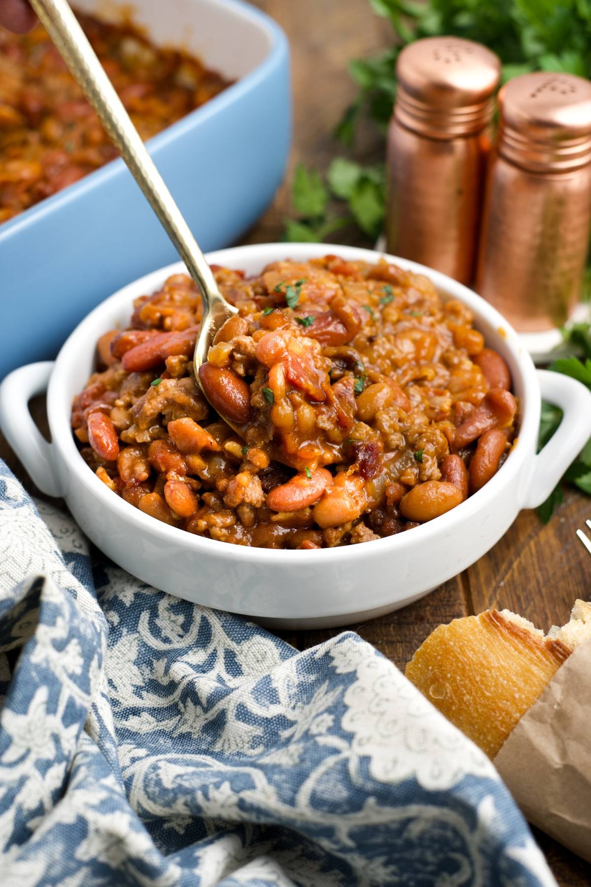 A white bowl of cowboy baked beans with a spoon, next to bread, herbs, and salt and pepper shakers.