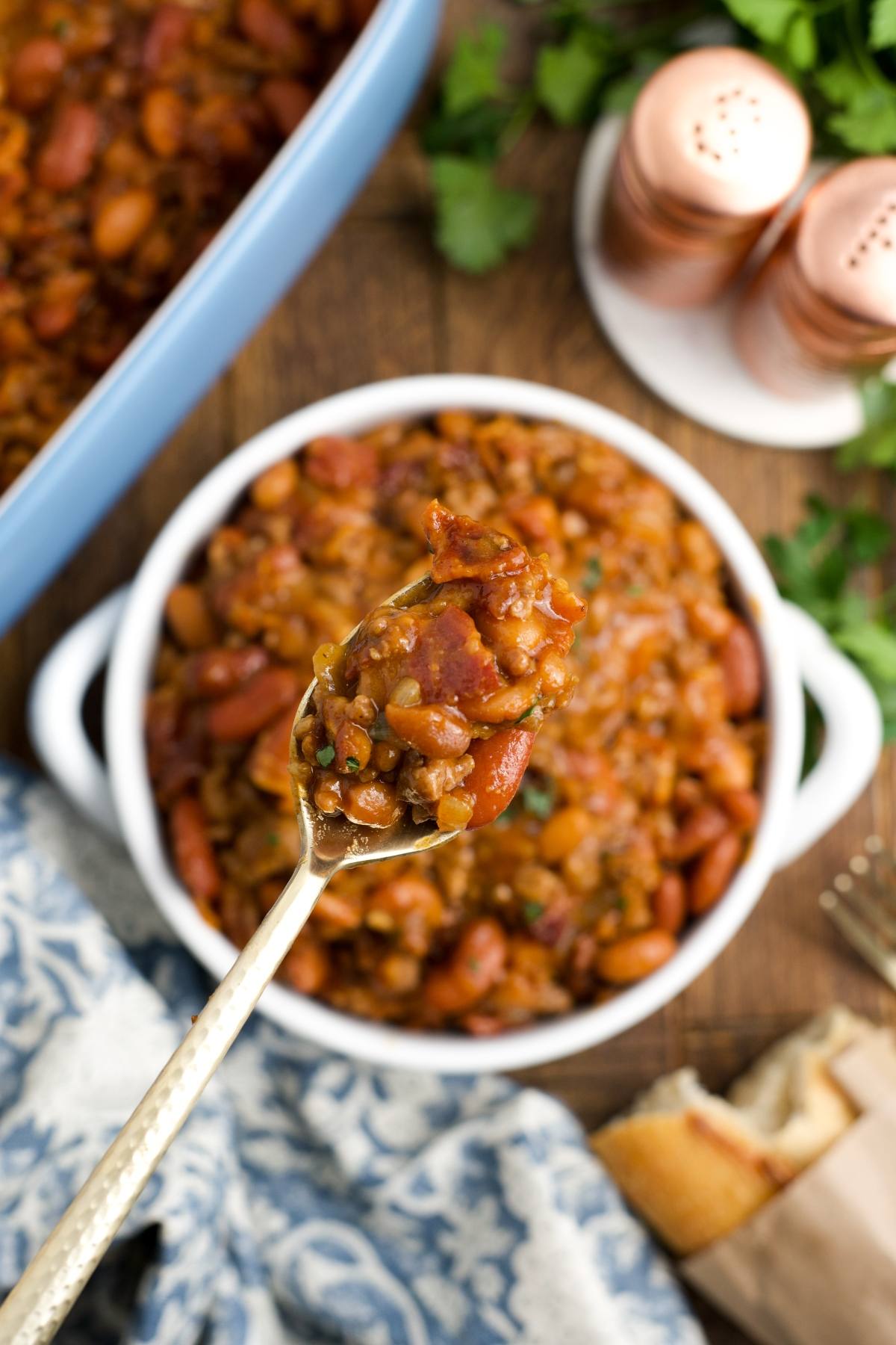 A spoonful of cowboy baked beans held above a dish, with bread and salt shakers in the background.