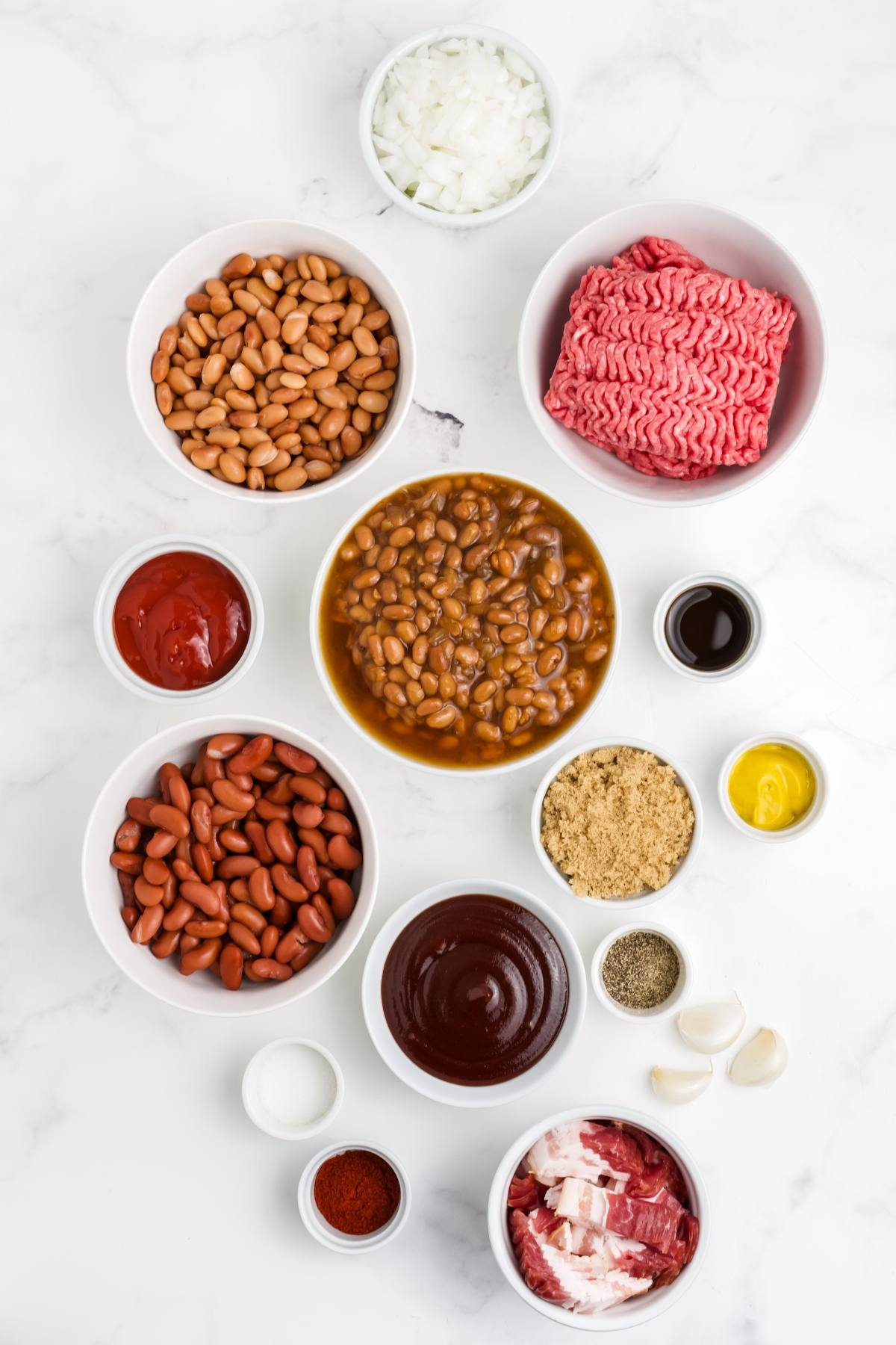 Various bowls with beans, ground beef, seasonings, onions, ketchup, and barbecue sauce on a white surface.