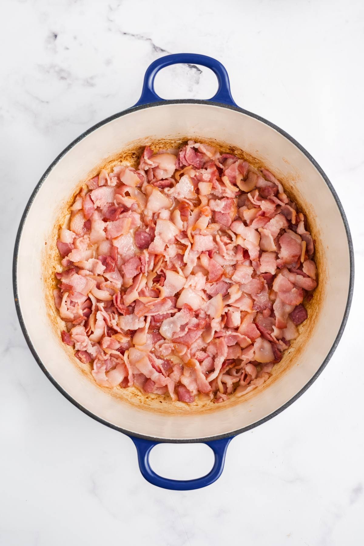Chopped cooked bacon pieces in a large blue-handled pot on a white marble surface.
