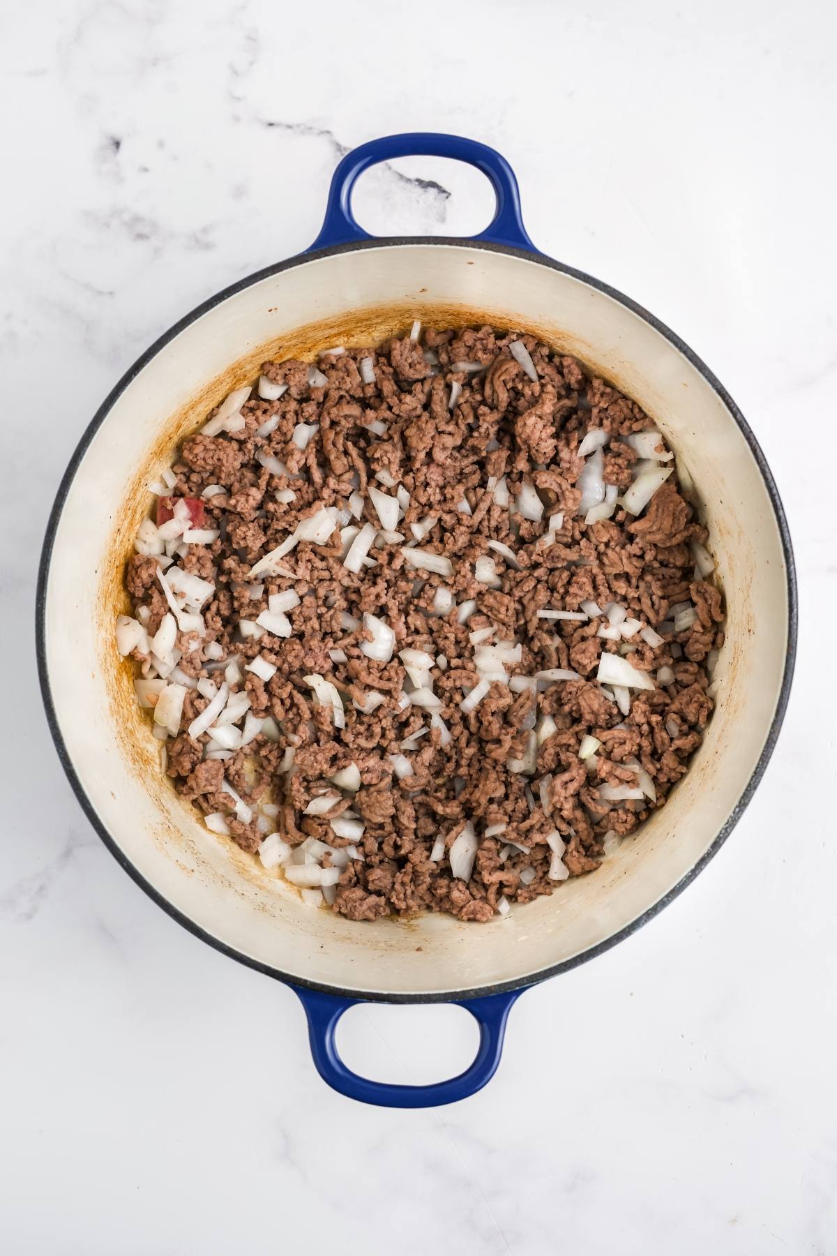 Ground beef and chopped onions cooking in a large blue Dutch oven on a white marble surface.