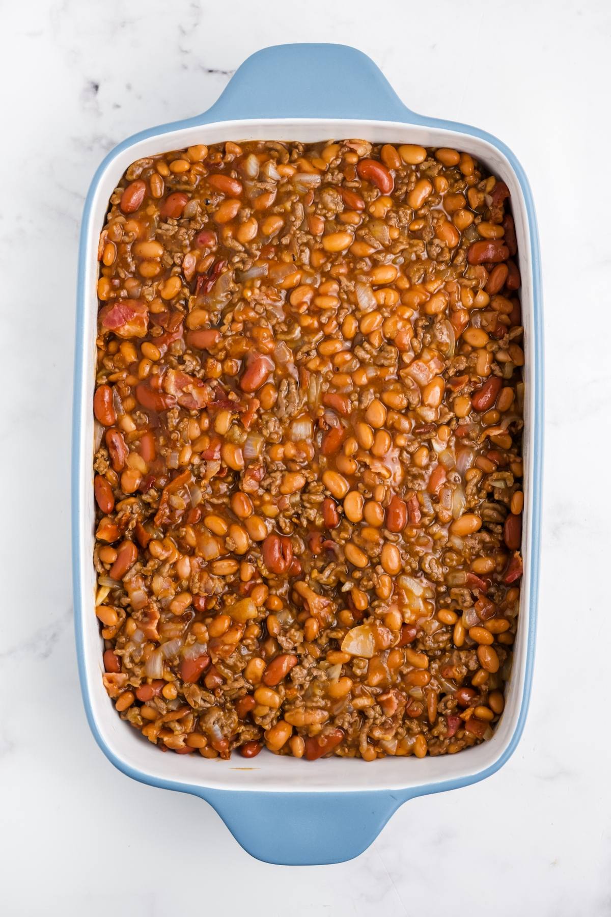 A blue baking dish filled with baked beans mixed with ground beef, onions, and tomato sauce.