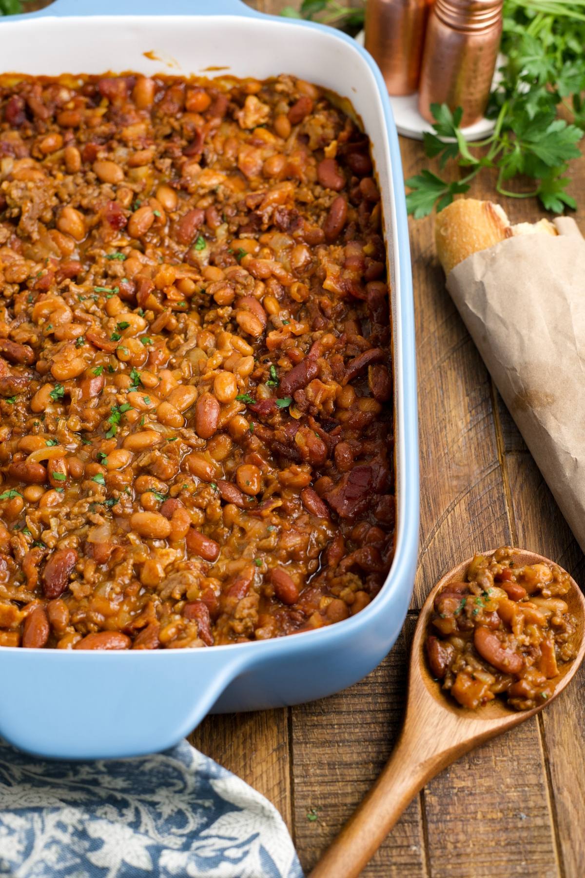 cowboy Baked beans with ground meat in a blue dish, with a wooden spoon of beans and bread on a wooden table.