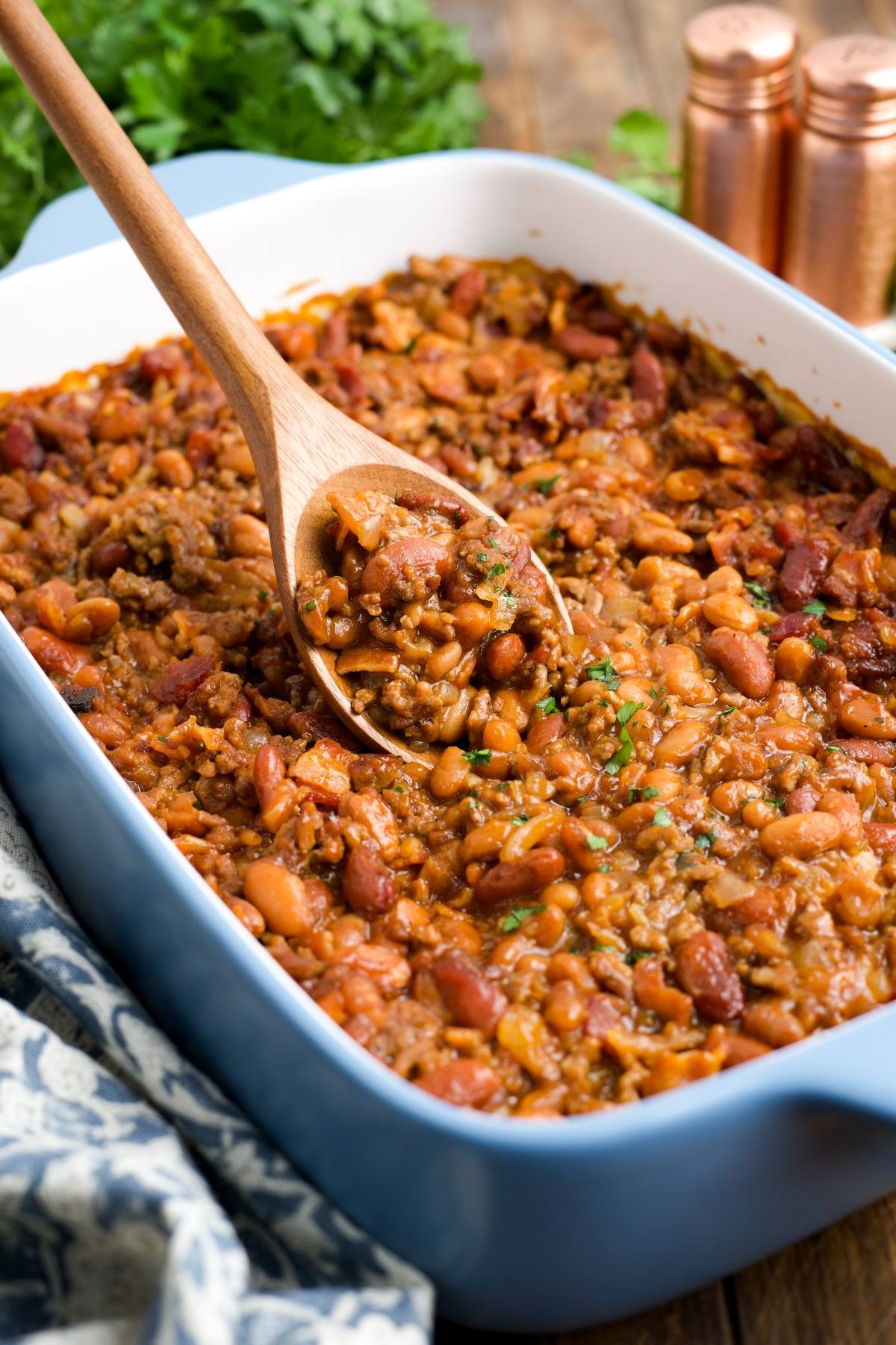 A wooden spoon scoops hearty cowboy baked beans from a blue casserole dish on a wooden table.