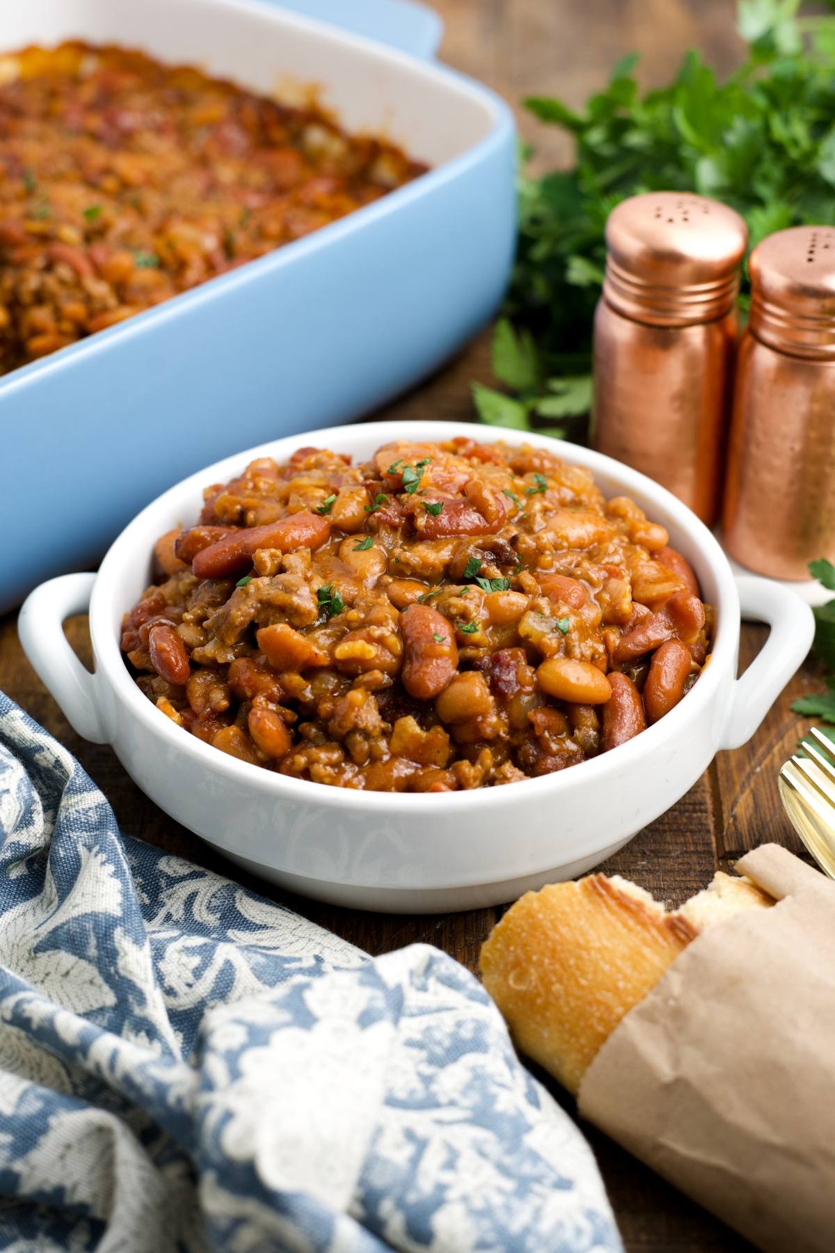 A bowl of cowboy baked beans with ground meat, garnished with parsley, beside bread and copper salt and pepper shakers.