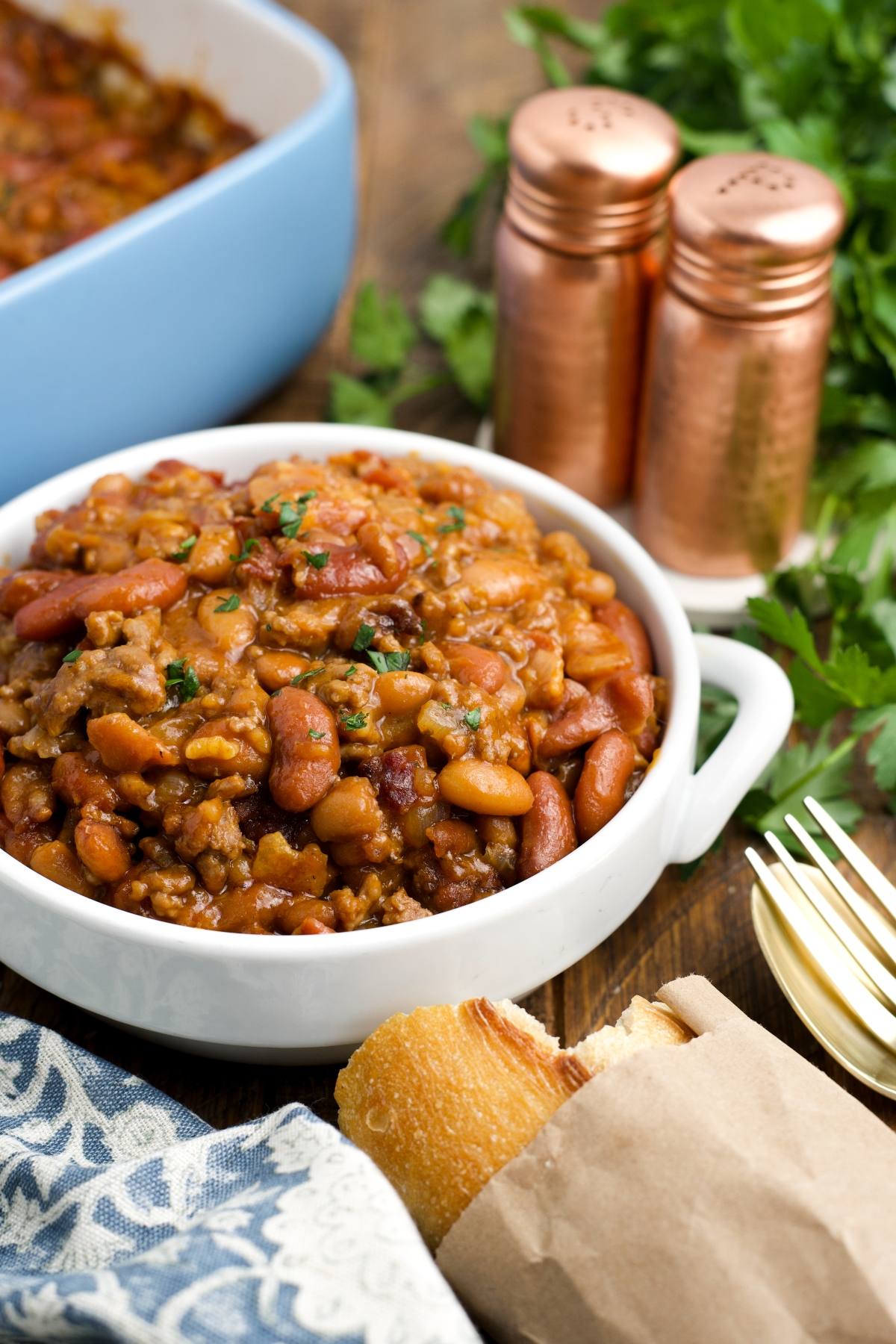 A bowl of cowboy baked beans with ground meat, served with bread, parsley, and salt and pepper shakers.