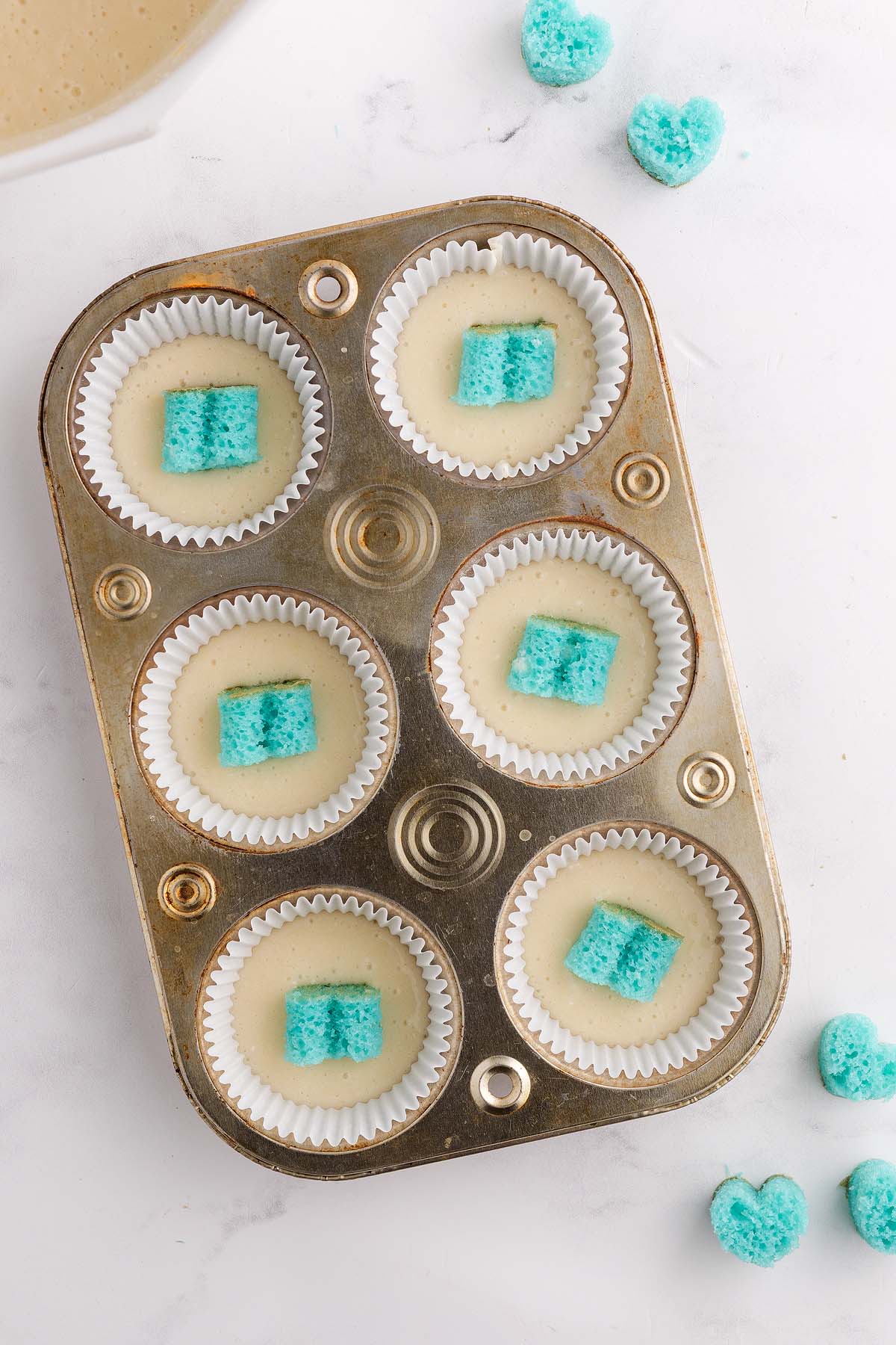 Muffin tin with cupcake liners, batter, and blue heart-shaped pieces inside each cup, perfect for making gender reveal cupcakes on a white surface.