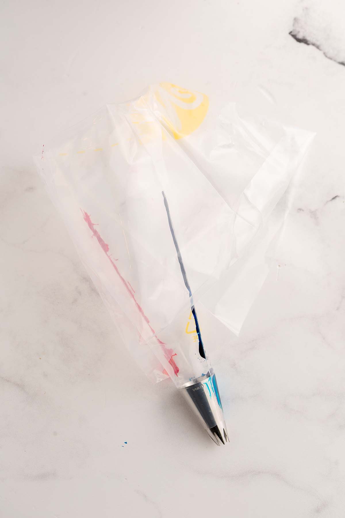 Piping bag filled with red, yellow, and blue frosting, fitted with a metal tip, on a white surface&mdash;perfect for decorating gender reveal cupcakes.