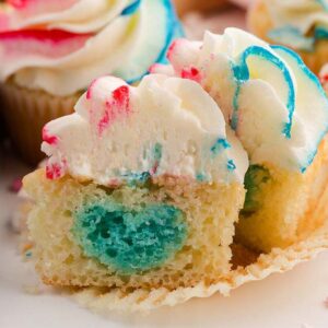 A gender reveal cupcake with colorful heart-shaped filling, topped with swirled frosting, cut open to show the festive surprise inside.