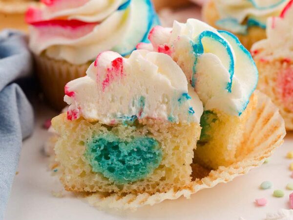 A gender reveal cupcake with colorful heart-shaped filling, topped with swirled frosting, cut open to show the festive surprise inside.