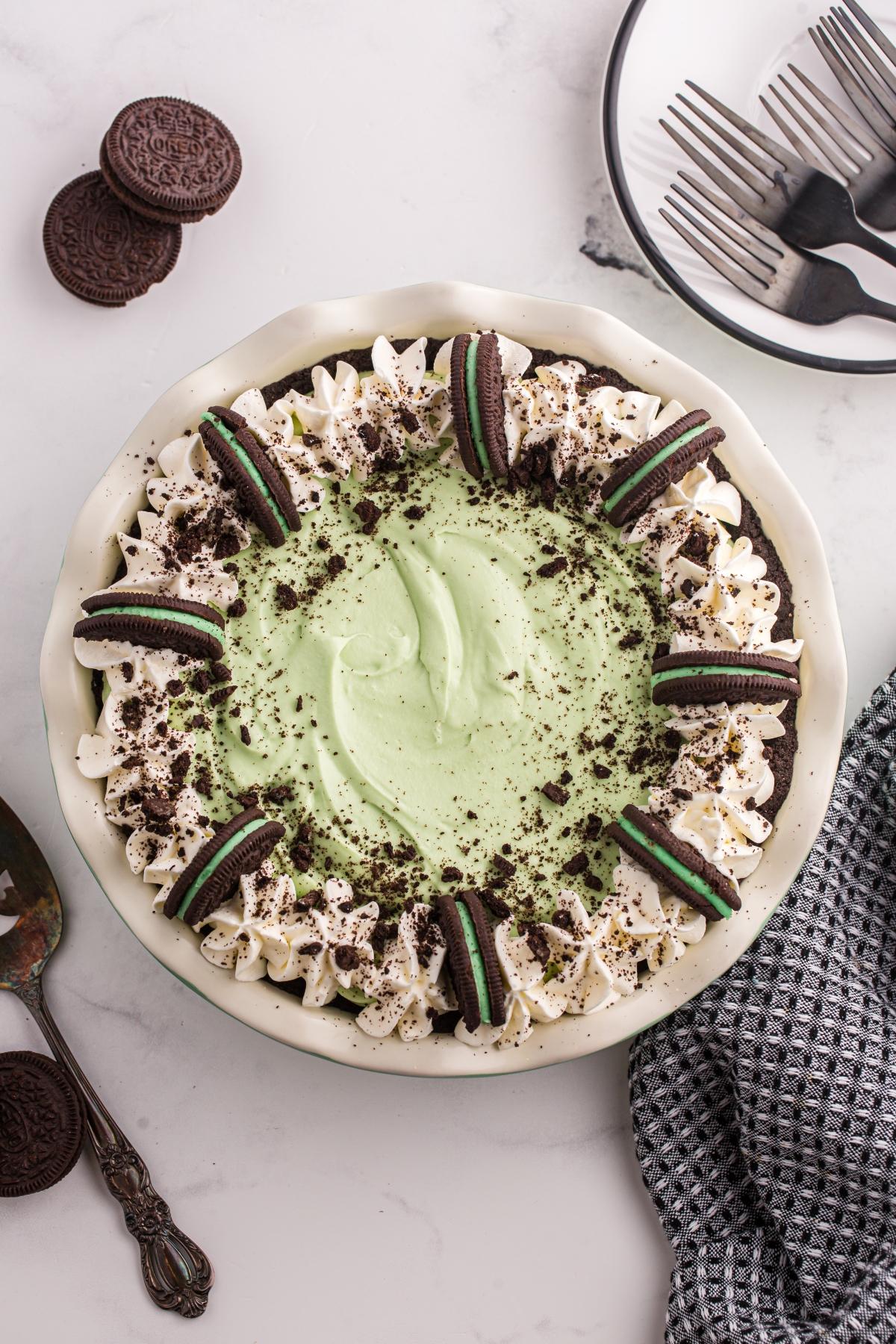 A grasshopper pie topped with whipped cream, Oreo halves, and cookie crumbs on a white marble surface.