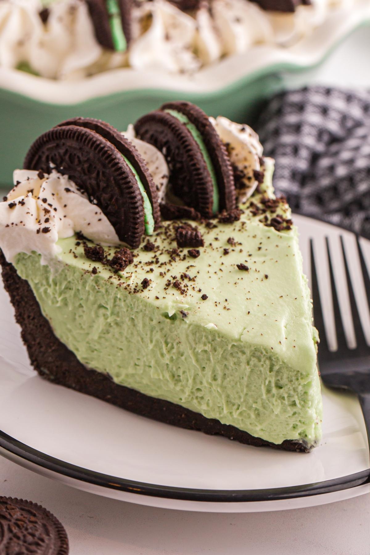 A slice of grasshopper pie topped with whipped cream and Oreo cookies on a white plate with a fork.