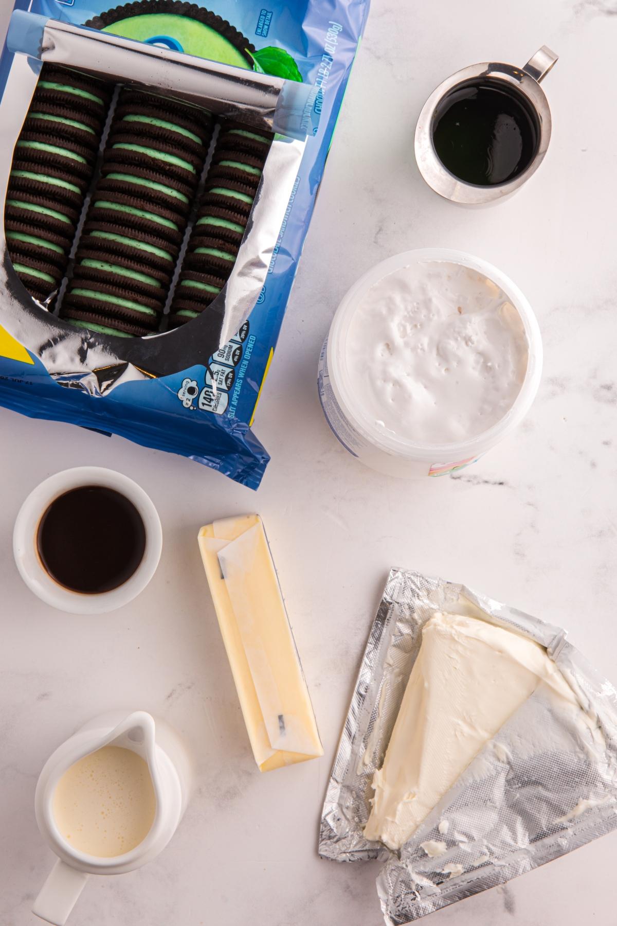 Top view of cookies, cream cheese, butter, coffee, cream, chocolate syrup, and whipped topping on a white surface.