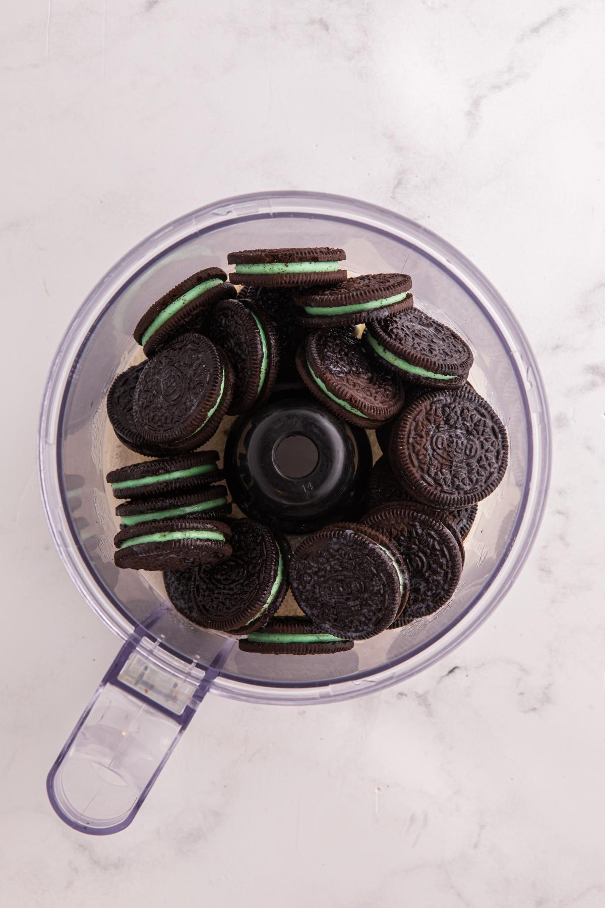 A food processor bowl filled with chocolate sandwich cookies with green cream filling, on a marble countertop.